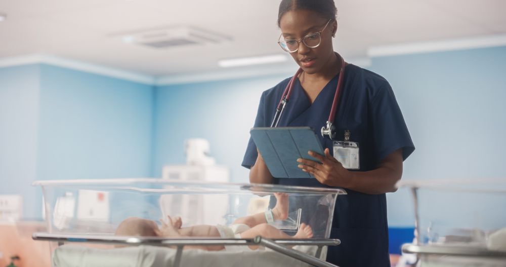 Young Black Nurse Using Tablet Computer Checking Up on a Newborn Baby in Maternity Ward Facility.