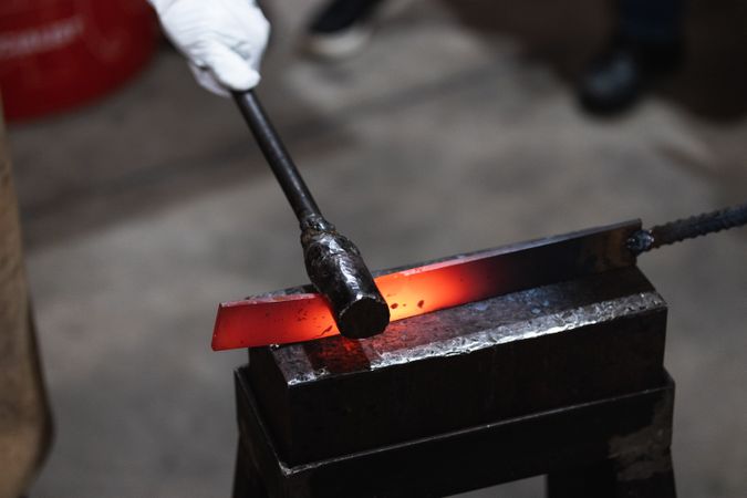 Blacksmith in gloves hot hitting metal part on anvil