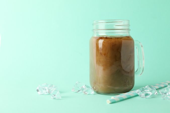 Jar of ice coffee, ice cubes and straws on mint background