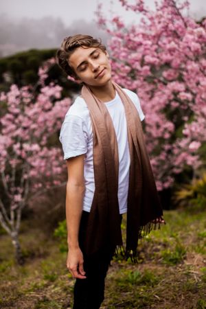 Young man in brown scarf standing beside pink flower tree outdoor