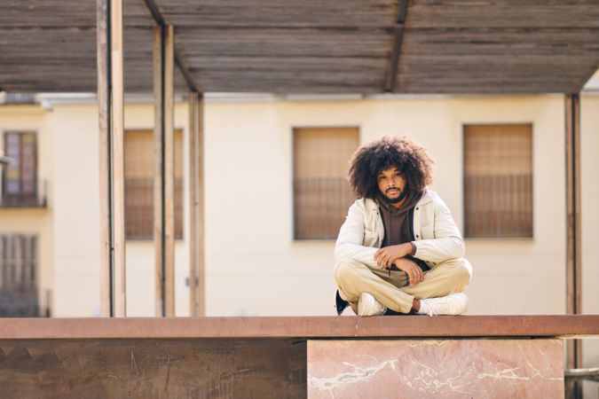 Young man with afro hair sitting cross-legged on a step on a quiet city street looking at the camera