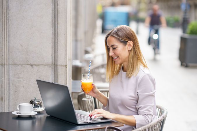 Woman in grey sitting in cafe outside with laptop and juice