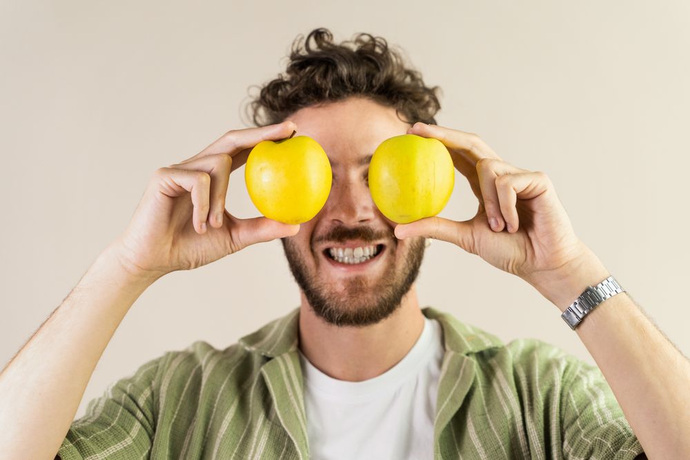 Funny young man holding two fresh apples over his eyes.