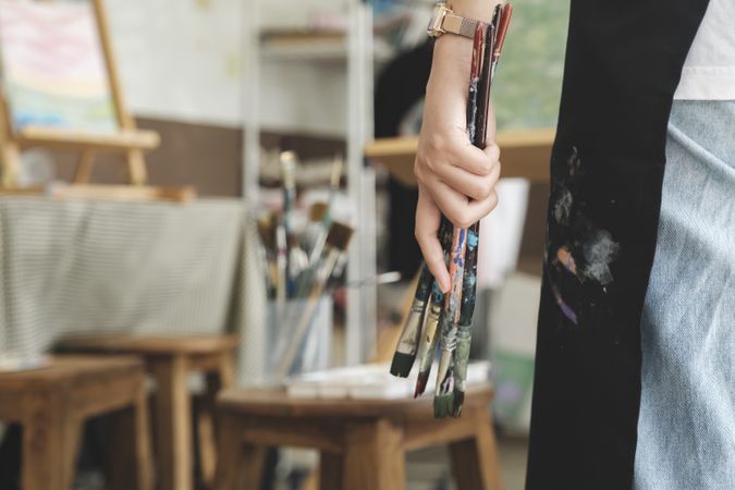 A close-up of the artist hands wearing an apron smeared with paint. Clutching many brushes and paintbrushes.