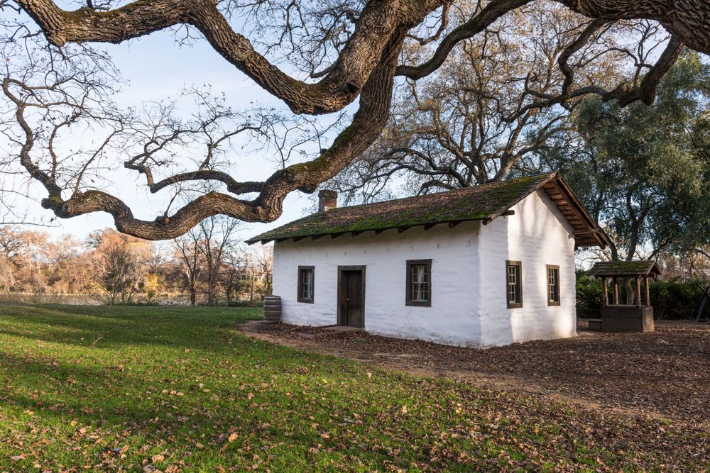 Small cream adobe house under large oak trees in California