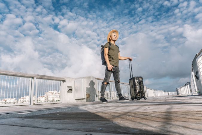 Full length side view serene tourist holding luggage while stands in a brown hat and ripped jeans