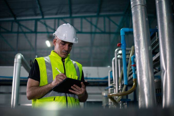 A male industrial engineer wearing a safety helmet and reflective vest is inspecting equipment in a factory. He is using a digital tablet to review data. Background show industrial pipes and machinery