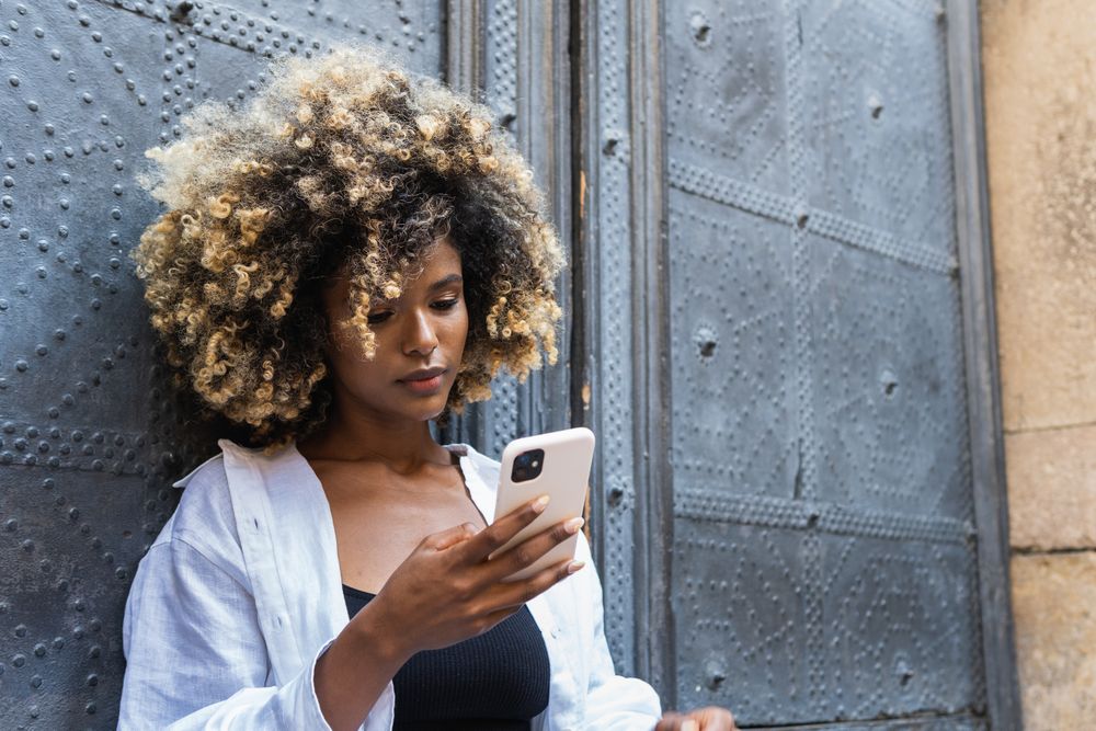 Young Black woman speaking on smartphone while sitting near building