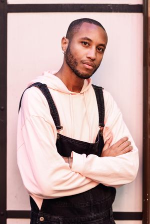 Male in denim overalls and earrings with arms crossed against a pink wall