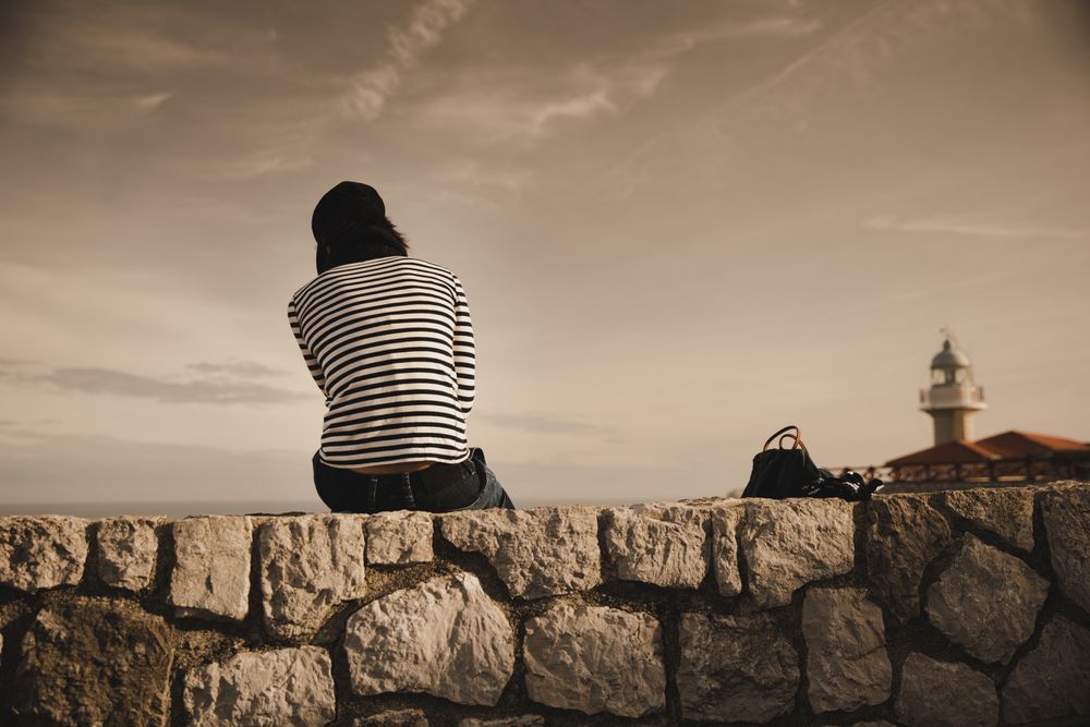 Back view of elegant woman in cap sitting on meadow near beacon on shore and wonderful sky