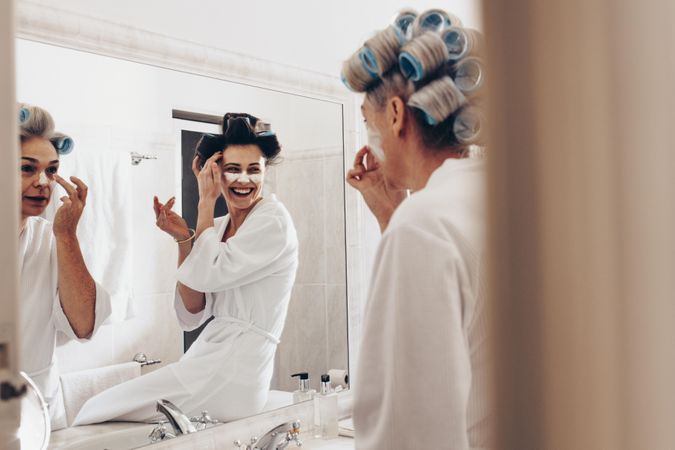 Two women in bathrobes applying cream on face standing in front of mirror