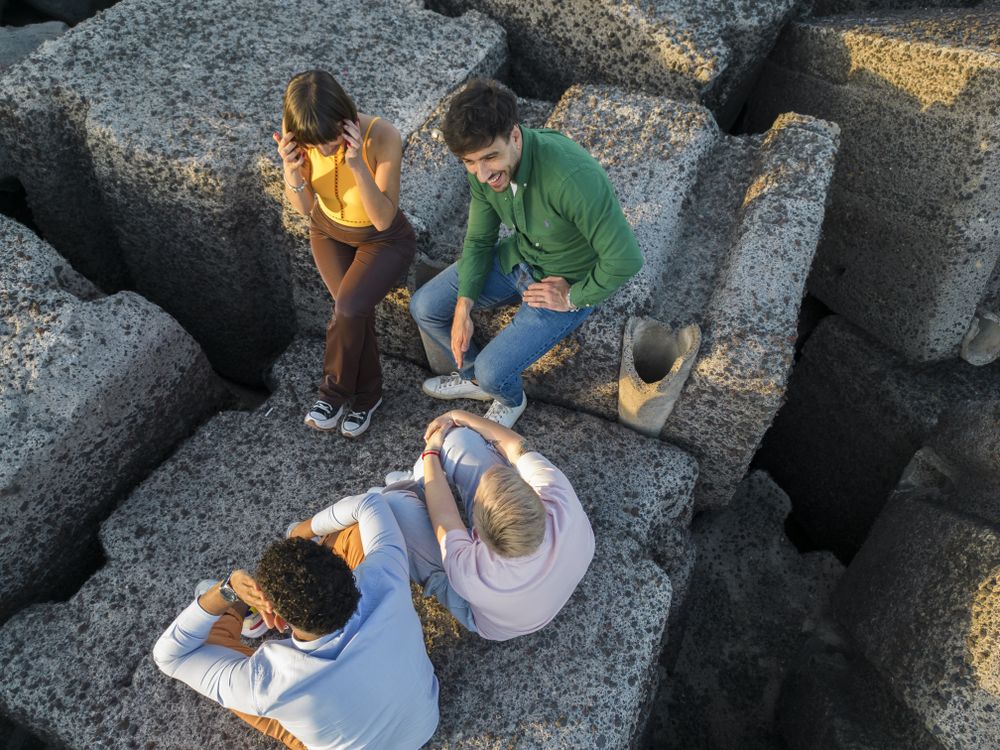 Aerial view of diverse friends sitting on rocks by the sea at sunset