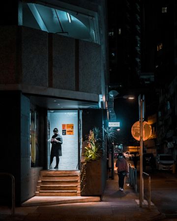 Person walking on sidewalk while woman standing at doorstep of a building at night in Hong Kong