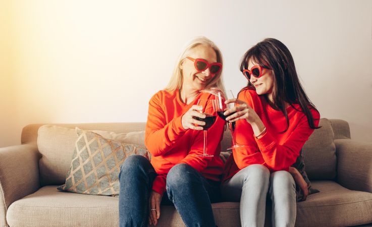 Adult daughter and mother in matching shirts and sunglasses toasting with wine at home