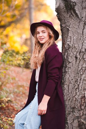 Smiling teenage girl in purple coat and hat leaning on tree