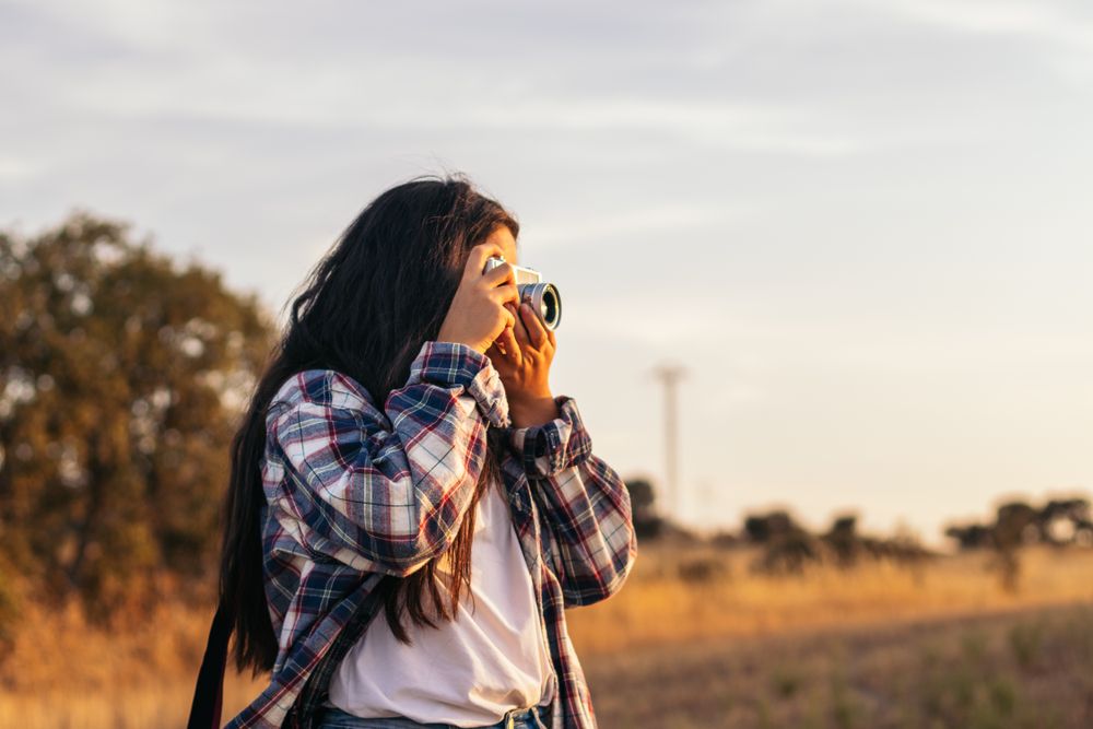 Young woman taking photos with analog camera in the field at sunset