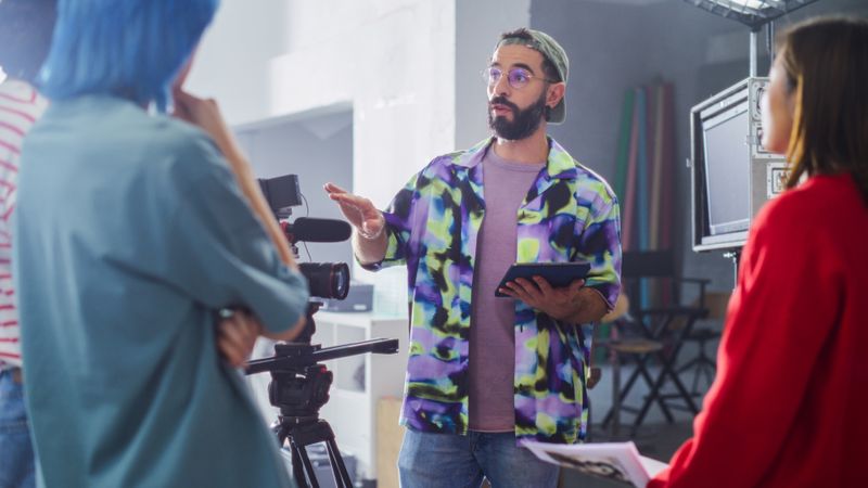 Young Male Director With Beard And Glasses, Instructing Diverse Team On Film Set, Holding Tablet, Surrounded By Camera Equipment And Crew In Creative Studio Environment.