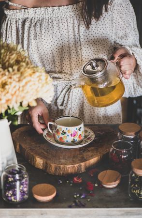Woman in pouring tea in floral cup and saucer from glass teapot, close up, vertical composition