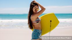 Portrait of a fit young woman on a beach carrying a bodyboard 0vyepb