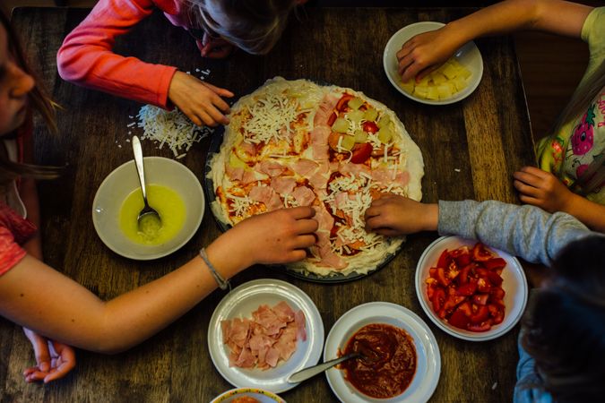 Top view of four people adding topping on a pizza dough