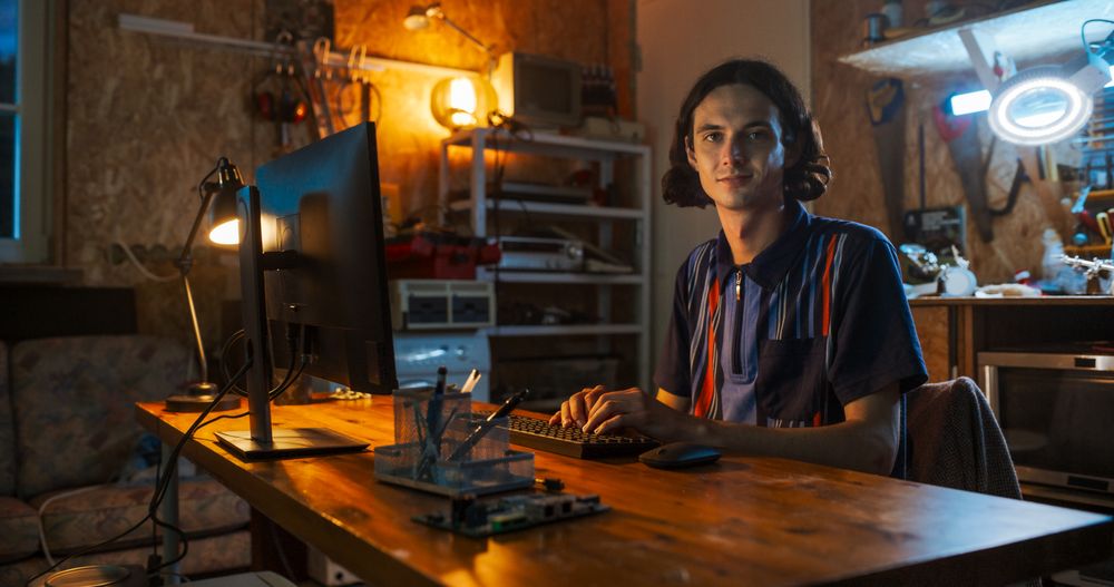 Portrait of White Male Software Engineer Programming On Desktop Computer In Home Garage, Looking At Camera And Smiling.