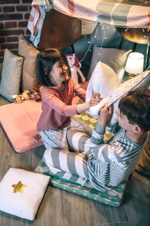 Excited boy and girl laughing in cushions battle on diy teepee in house