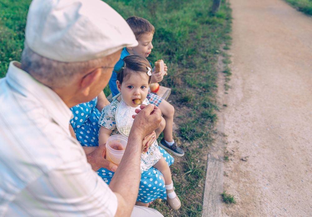 Back of older man feeding baby girl sitting in a bench