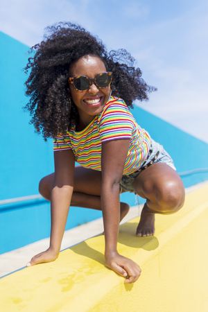 Smiling Black woman with sunglasses crouched down on yellow wall while looking to camera
