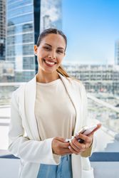 An entrepreneur woman is smiling and holding a cell phone while she looks at the camera. She is wearing a white jacket and jeans. Concept of entrepreneurism and modernity.