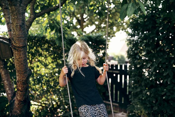 Blonde girl with braided hair standing on outdoor swing while looking down in backyard