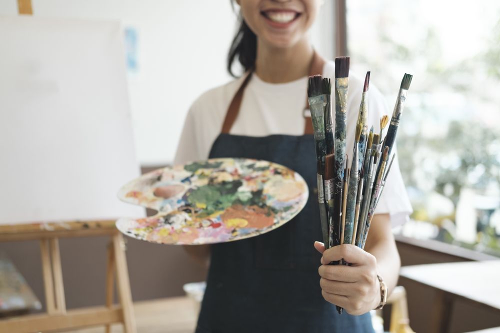 A close-up of the artist hands wearing an apron smeared with paint. Clutching many brushes and paintbrushes.