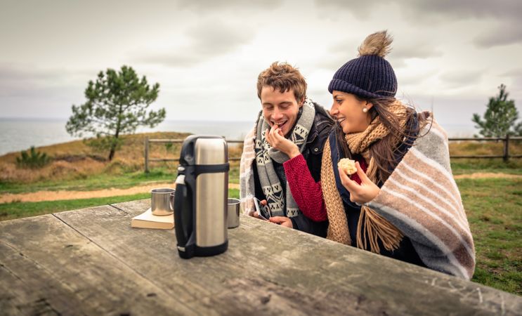 Couple wrapped in blanket on cold park bench with coffee flask sharing muffin