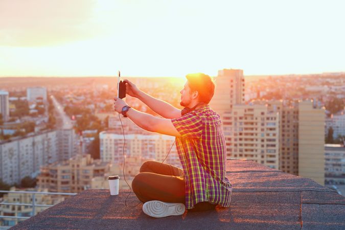Male sitting on roof taking selfie with tablet at sunset