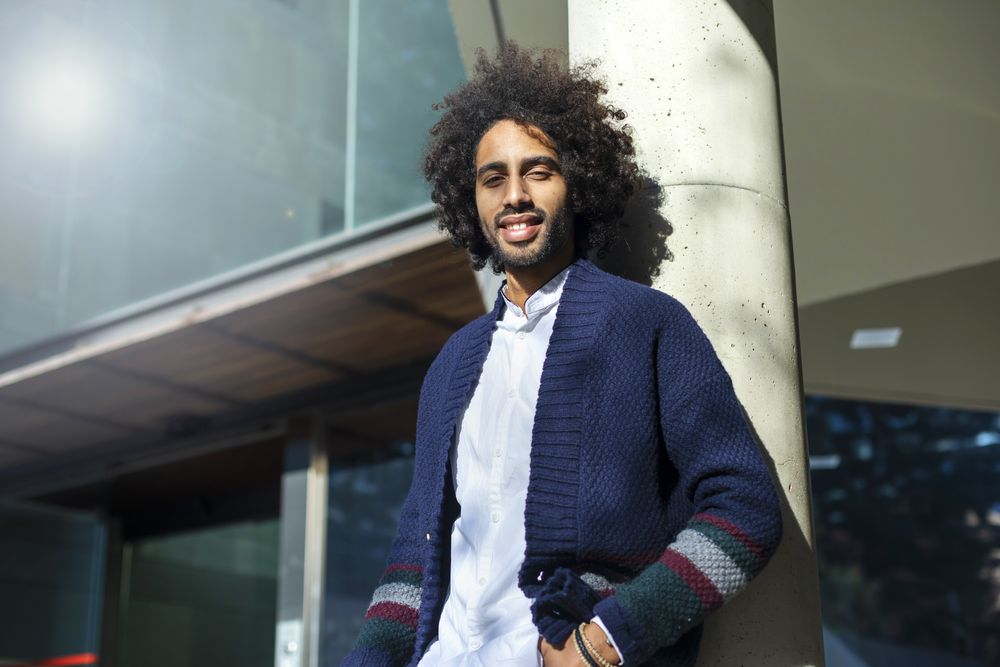 Portrait of Black man in cardigan and jeans leaning on pillar outside on sunny day
