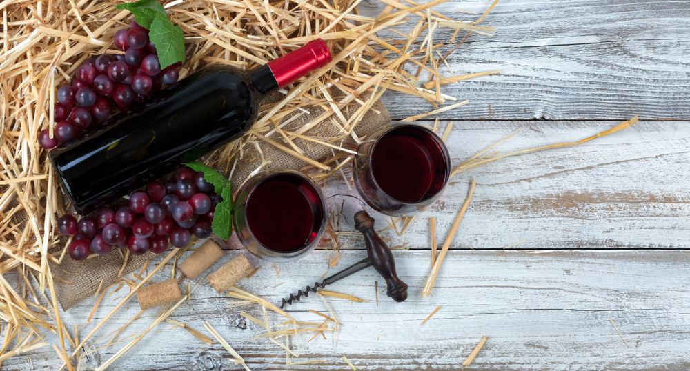 Bottle of red wine plus drinking glasses on top of straw and burlap with rustic wooden boards underneath