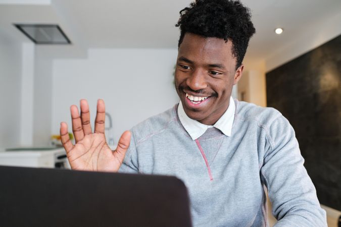 Cheerful Black man showing hi gesture against laptop at home