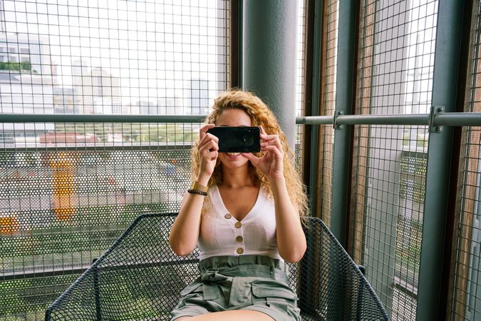 Pleasant woman taking photo with smartphone sitting in metal chair