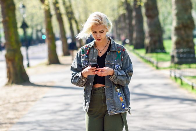 Blonde woman in jean jacket checking messages on cell phone while walking in park