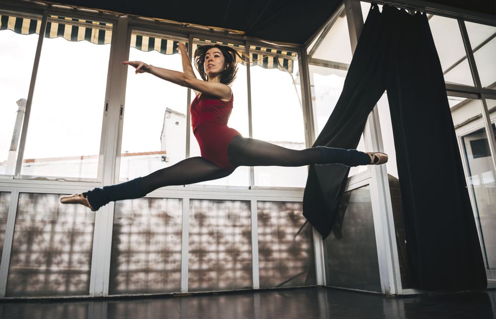 Young slim ballerina jumping above ground in studio flexing legs.