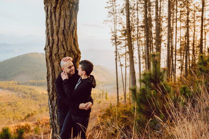 Smiling men hugging near tree in forest on hill