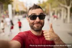 A young, bearded man, wearing sunglasses, smiles at the camera, while taking a selfie with his cell phone during a walk in the city bE9926