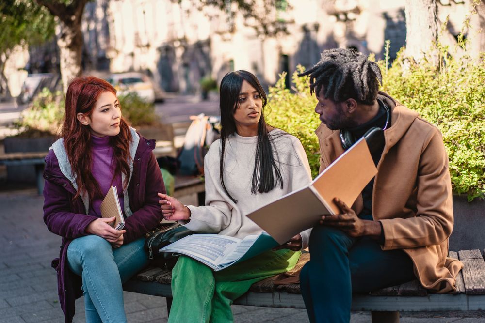 Three diverse students are deeply engaged in a group study session outside, sharing ideas and learning together