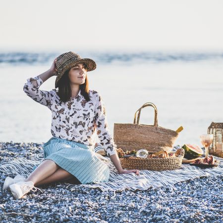 Young woman in hat sitting by the ocean with a picnic, watermelon, and wine, square crop