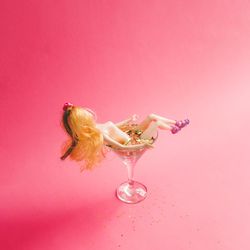 Doll bathing in martini glass full of gold glitter on pink background