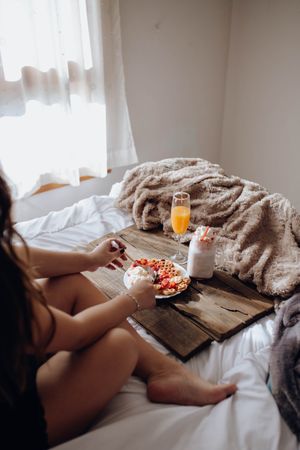 Young woman eating appetizing waffles and tasty drinks on tray on bed