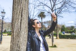 Side view of a young laughing hipster woman standing and leaning on a tree at park in sunny day while using a mobile phone to take a selfie