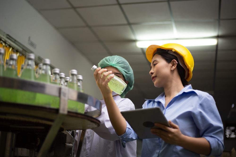 Asian female manager and worker holgin digital tablet checking information and quality control for juice drink product on bottling factory machinery.