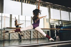 Back view of young slim ballerina jumping above ground in studio flexing legs.