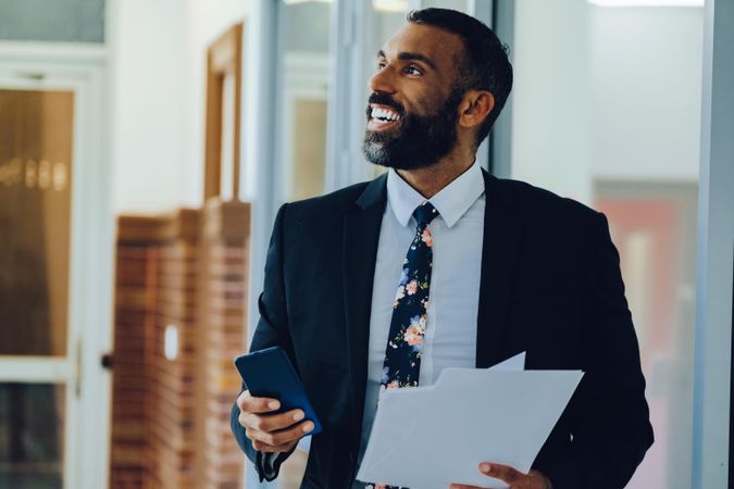 Happy businessman in suit and tie with papers standing in office while holding smart phone
