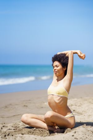 Woman in yellow swimwear stretching triceps above head on sandy beach